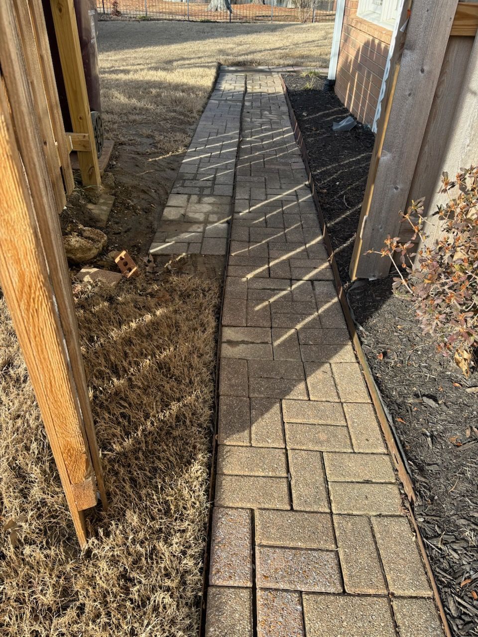 Brick pathway between a wooden fence and building; dry grass and mulch border the path.