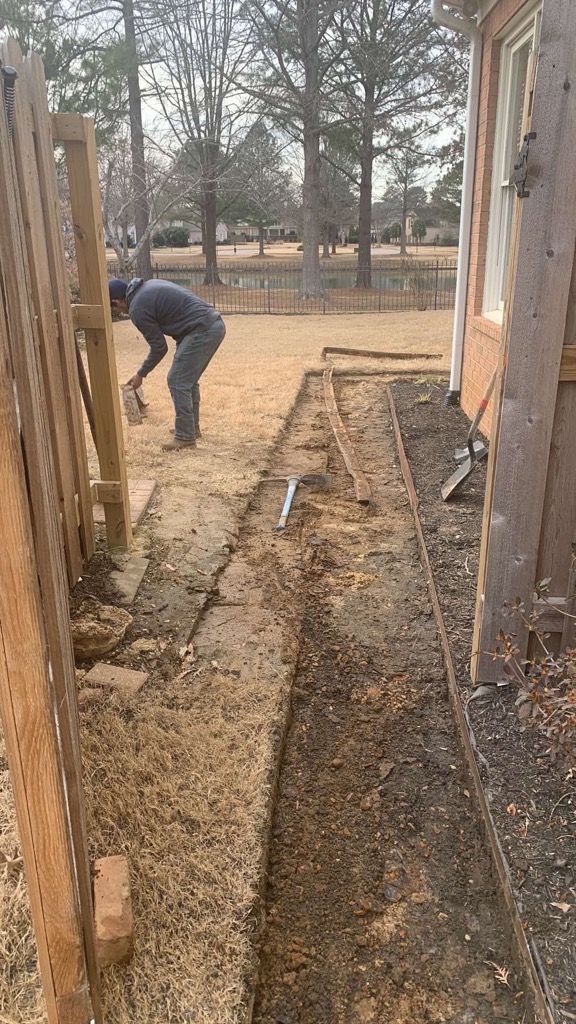 Man working on a narrow trench next to a house and fence. Dirt and tools are visible.