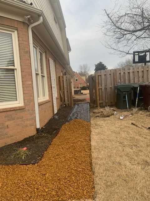 Pathway alongside a brick building, transitioning from mulch to gravel, leading to a wooden fence.