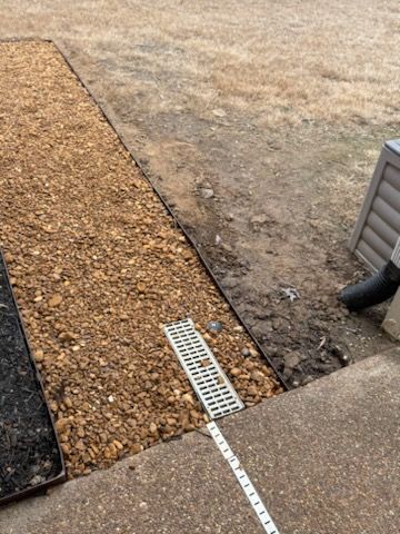 Gravel drainage bed with a grate. Brown gravel, concrete, dirt, black border, and a downspout are visible.