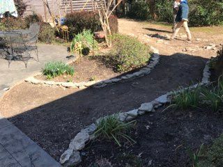 A person walks past a newly mulched, stone-edged garden path near a patio with a table and chairs.