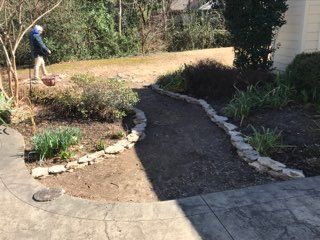 Man spreading material on a brown pathway edged with stones. Brown, unkempt yard. Outdoors.