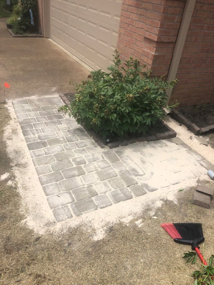 A gray paver walkway under construction, next to a bush and garage. Sand is spread around the pavers.