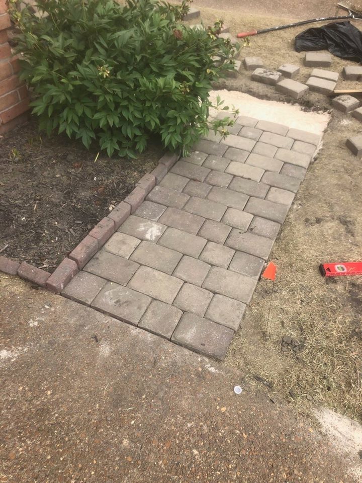 A brick walkway being constructed near a bush. Bricks are gray-brown; the setting is outdoors.