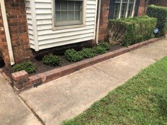 Brick-bordered flower bed with small green bushes and a concrete sidewalk in front of a house.