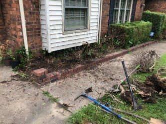 A brick-lined garden bed next to a brick house, with gardening tools scattered on the ground.
