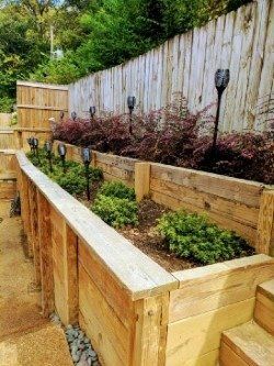 Wooden tiered garden beds with plants and solar lights against a wooden fence.