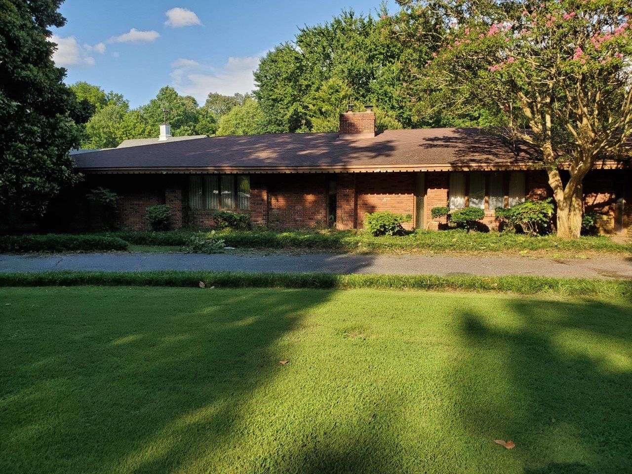 Low brick house with brown roof, surrounded by green grass, trees, and small shrubs under a blue sky.