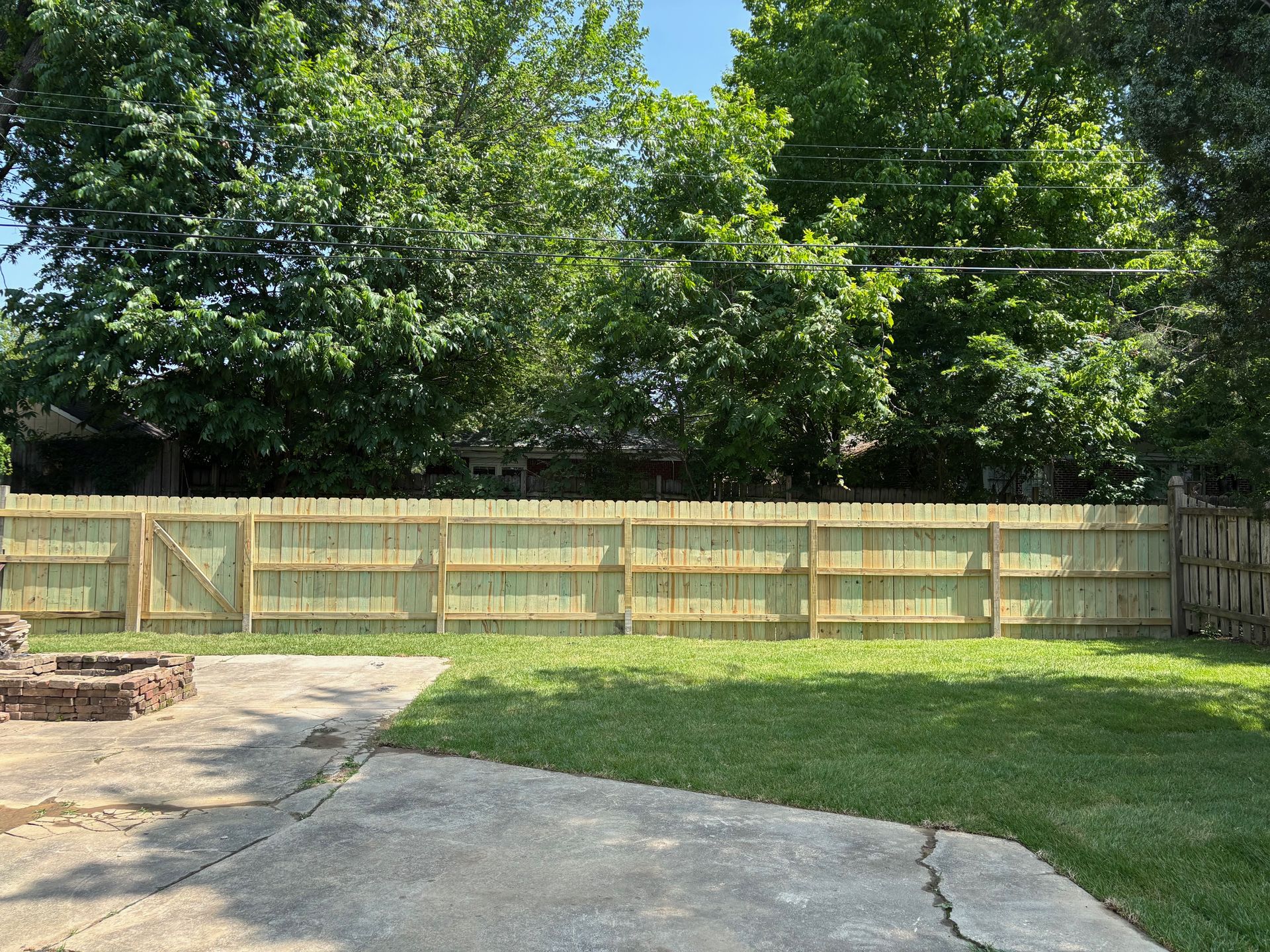 Wooden fence in a backyard with green grass and trees, and a concrete patio.