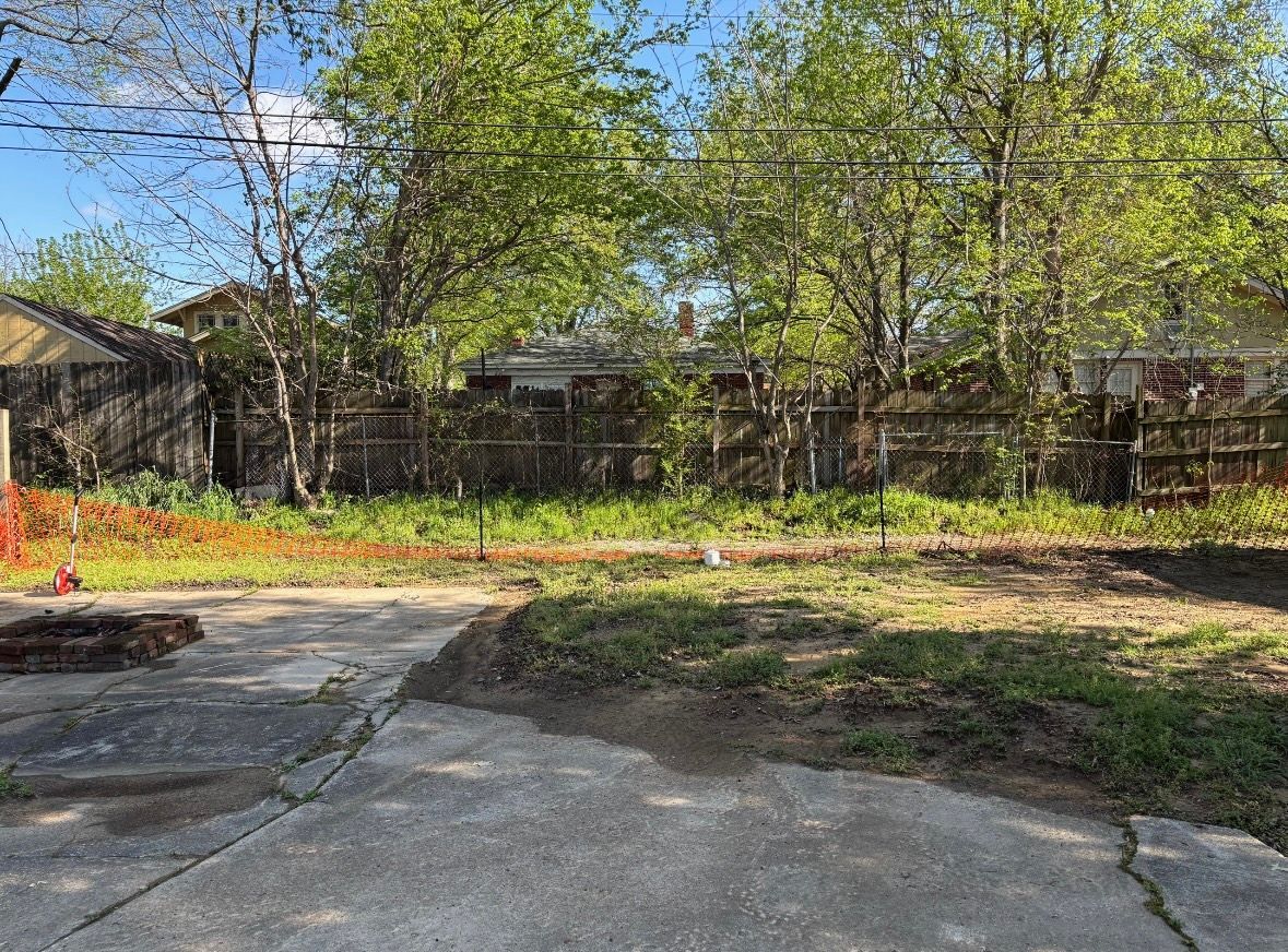 View of a backyard with trees, fence, and overgrown grass. A paved area is in the foreground.