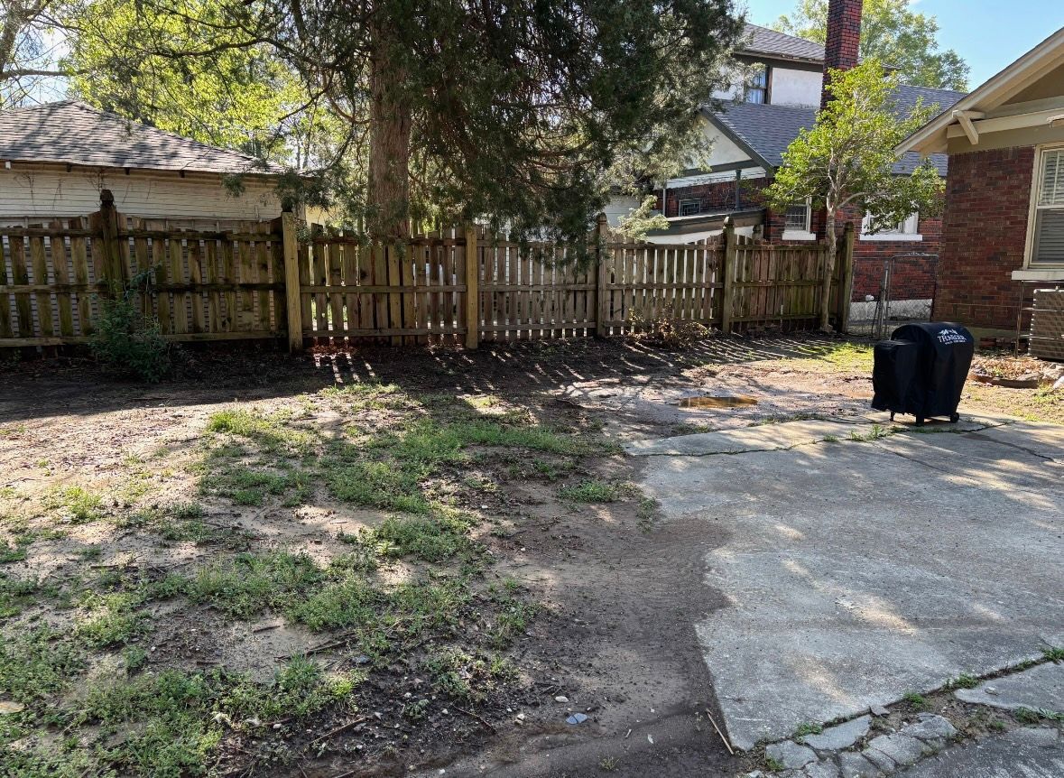 Backyard with fence, trees, and a concrete patio. Two black trash cans sit on the patio.