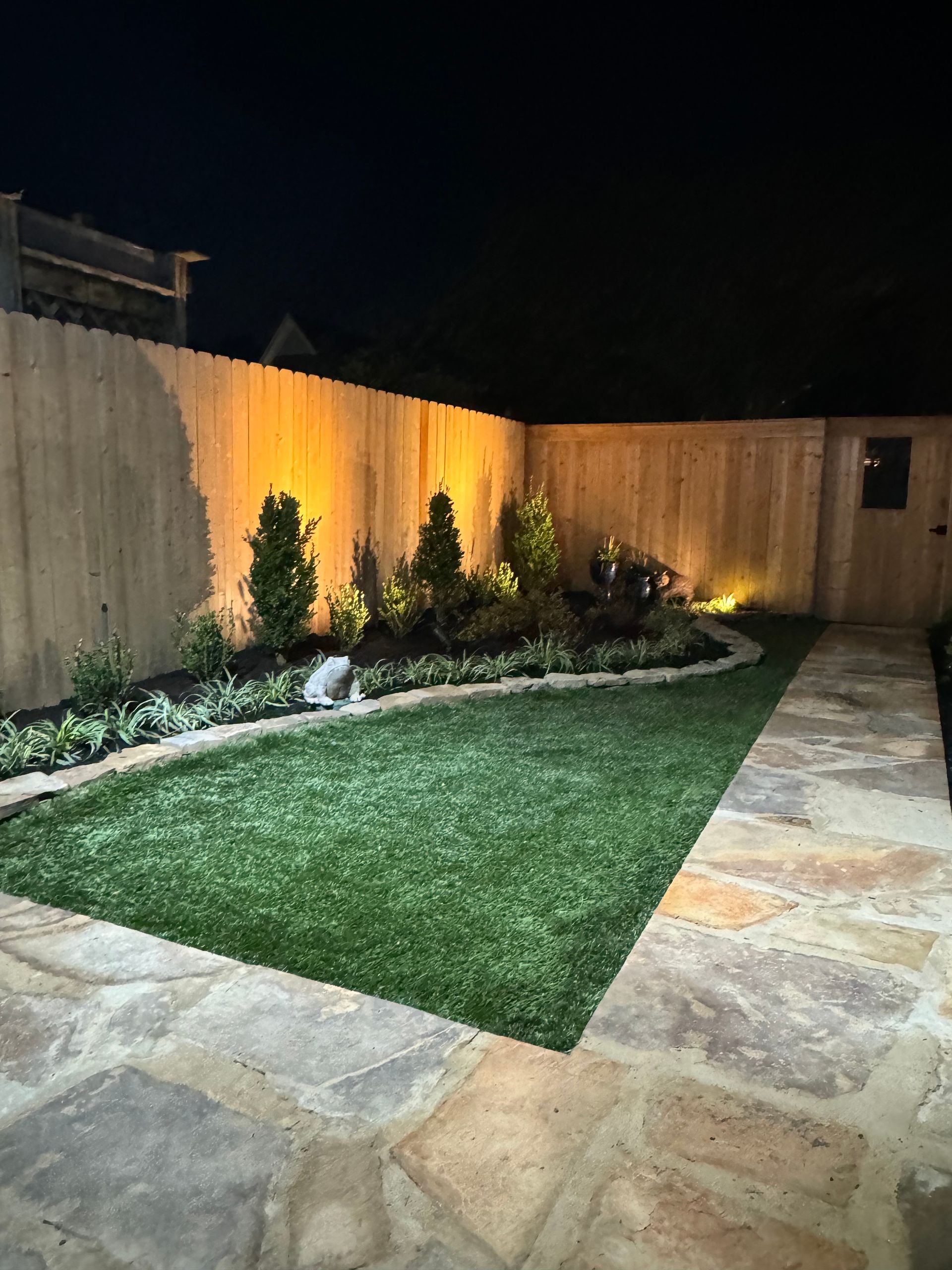 Backyard with artificial turf lawn, stone patio, lit garden, and wooden fence at night.