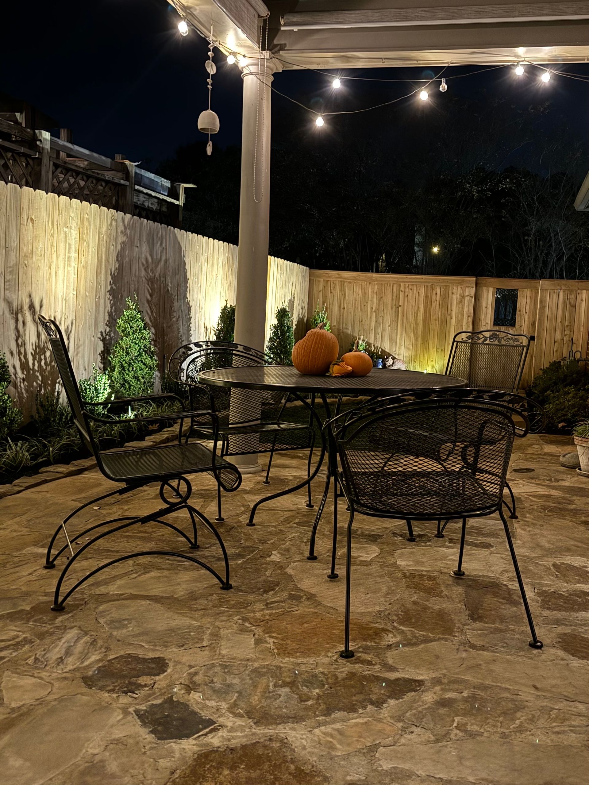 Patio at night with string lights, metal table and chairs, pumpkins, and a stone floor.