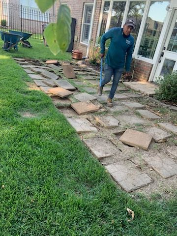 Man in teal shirt, jeans, and hat rebuilding stone pathway in a backyard. Grass and a brick house visible.