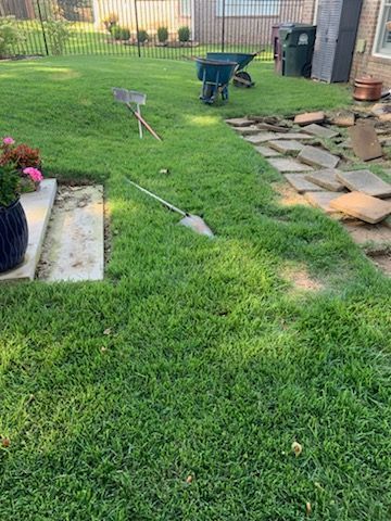 Lush green backyard with grass, stone path, tools, and a wheelbarrow. A blue flower pot sits on a concrete base.