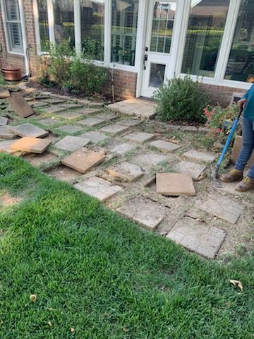 Stone pavers removed from a walkway in a grassy yard; a person holds a tool.