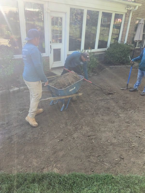 Three workers spread soil on a patio near a brick building. One pushes a wheelbarrow.