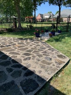 Stone patio in backyard, with potted flowers, grass, and a black fence.