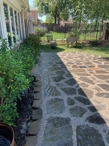 Stone path and patio in a backyard, bordered by bushes and a lawn, with a house on the left.