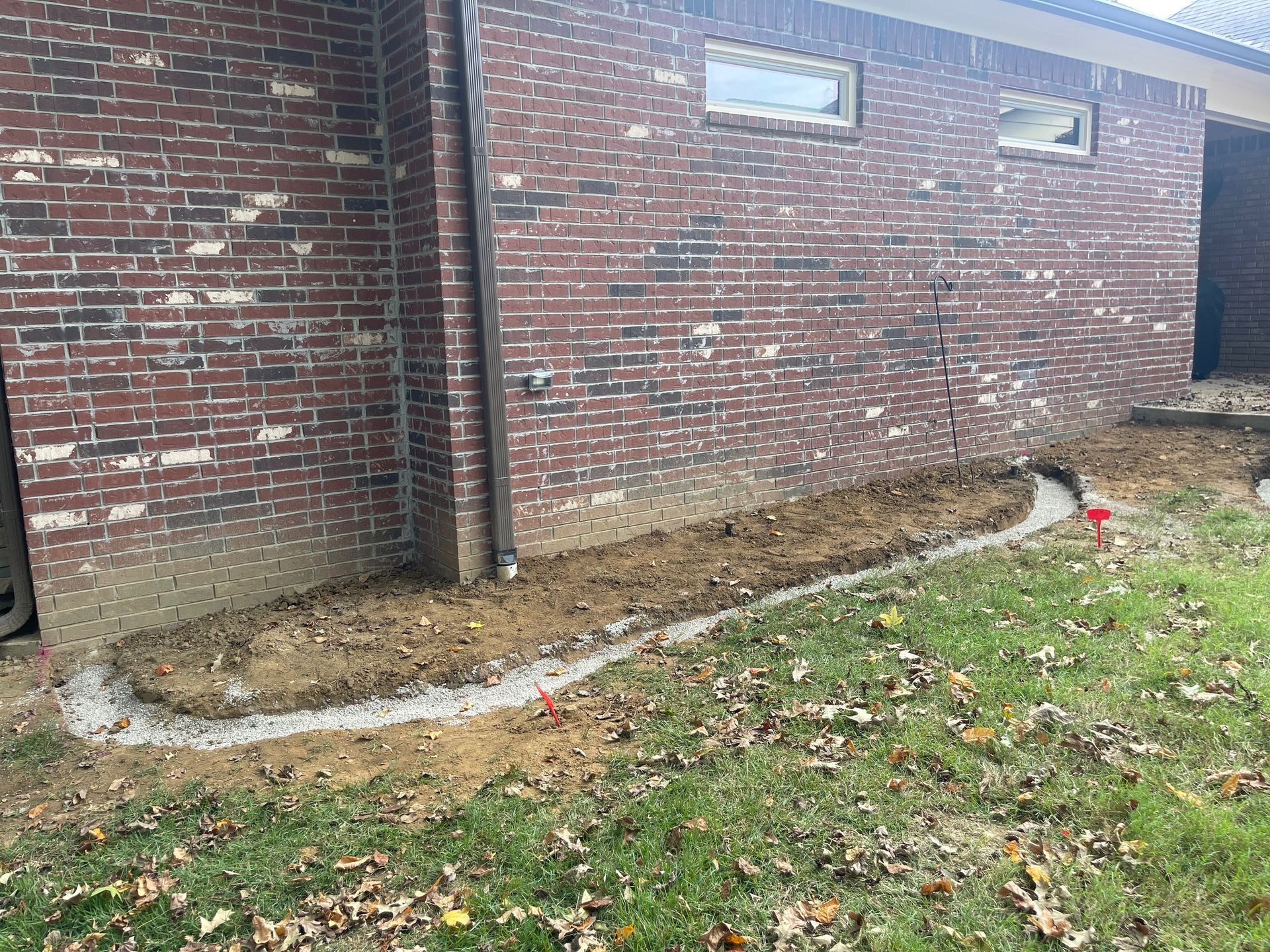 Brick building with a concrete border along the base; soil and grass are present.