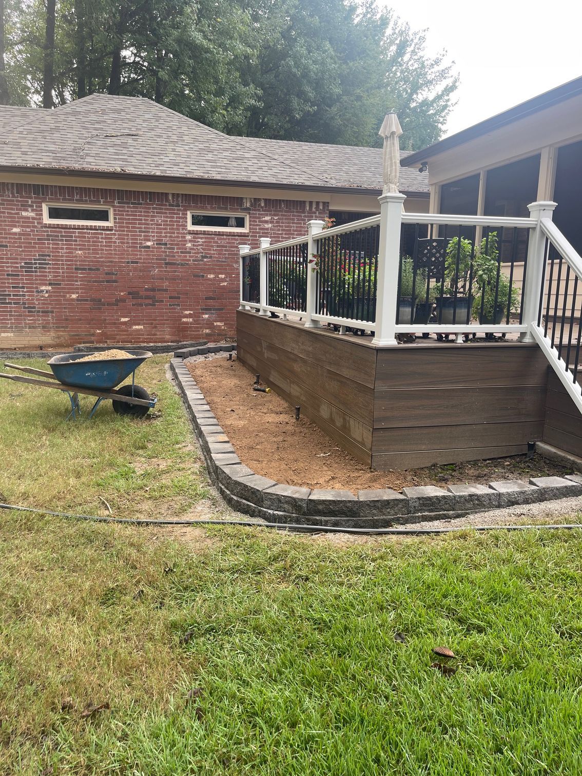 A backyard with a deck and garden bed lined with pavers; a wheelbarrow sits nearby.