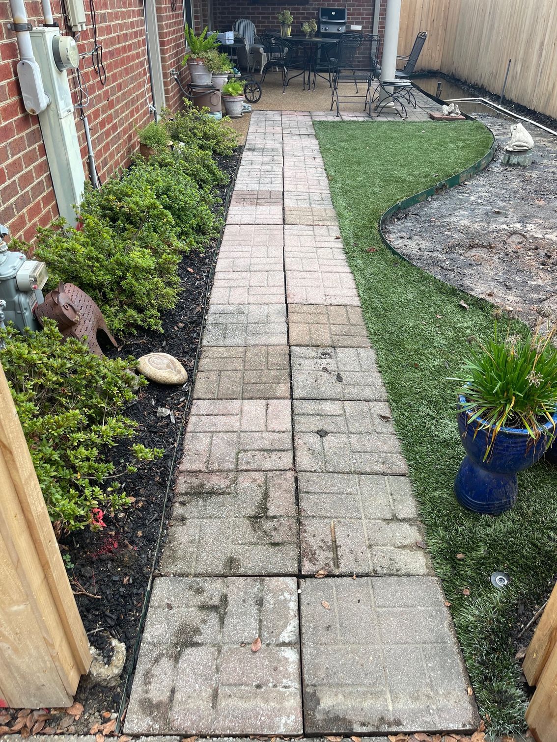 Brick walkway in a backyard, flanked by greenery and artificial turf.