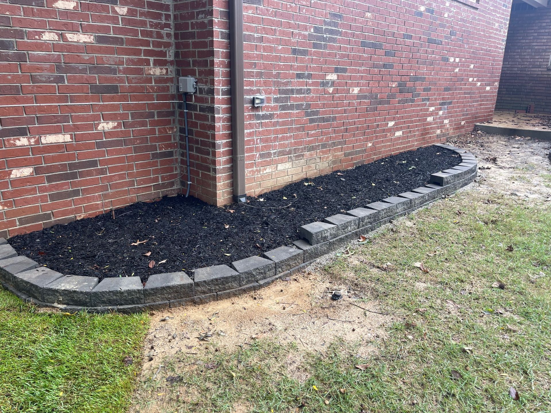 Black mulch in a brick-bordered flower bed against a red brick wall, on a grassy yard.