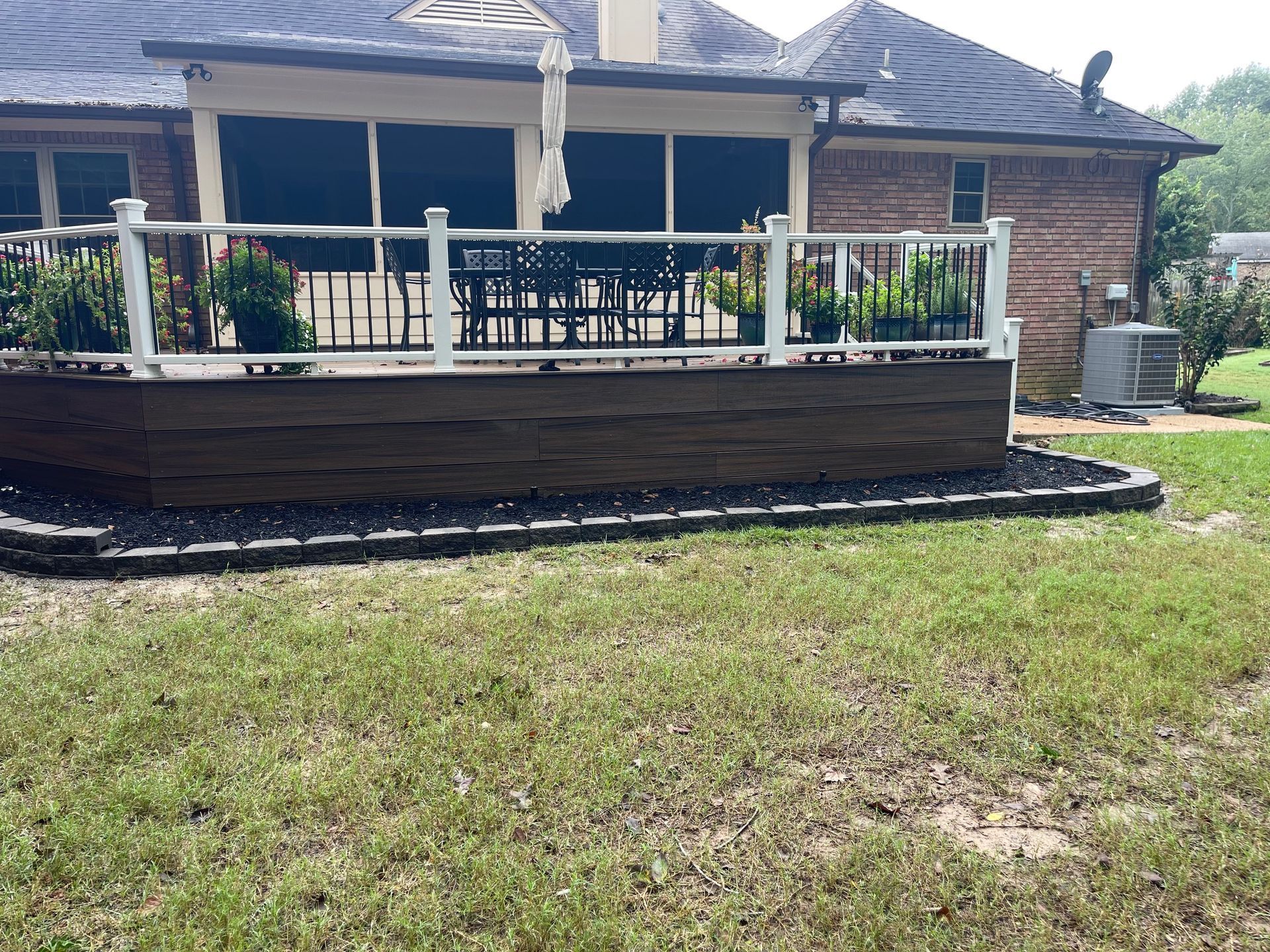 A backyard deck with a brown border, white railing, and black mulch border.