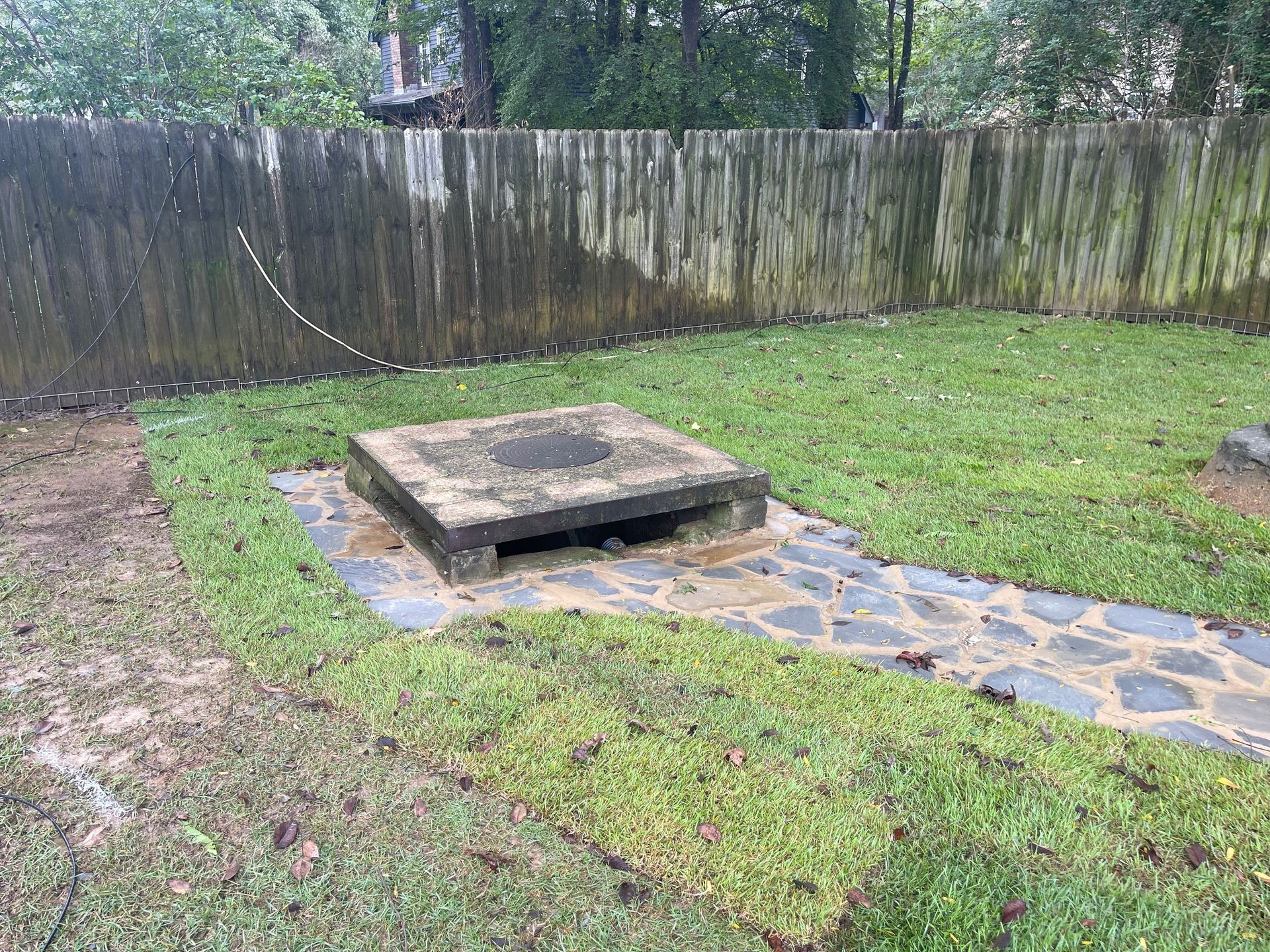 Concrete septic tank cover and stone path in a backyard, green grass, weathered wooden fence.