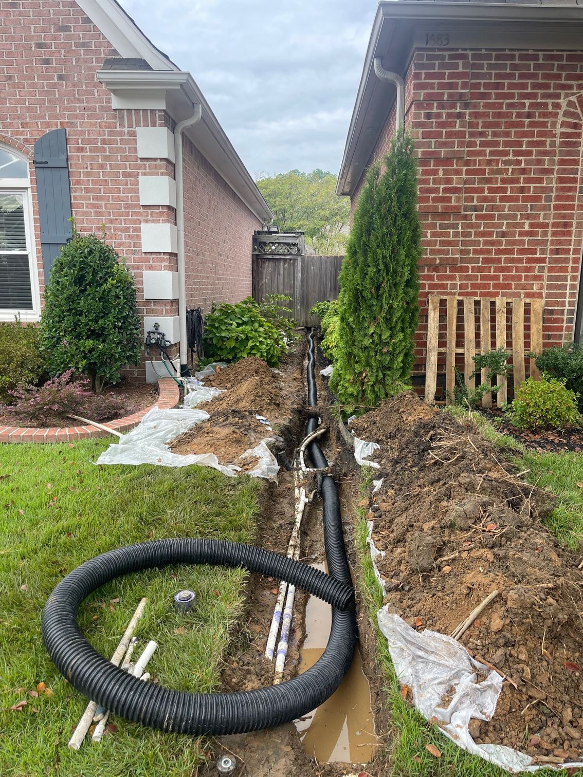 Trench being dug between two brick houses for a black drainage pipe. Grass, soil, and white PVC pipes are also visible.