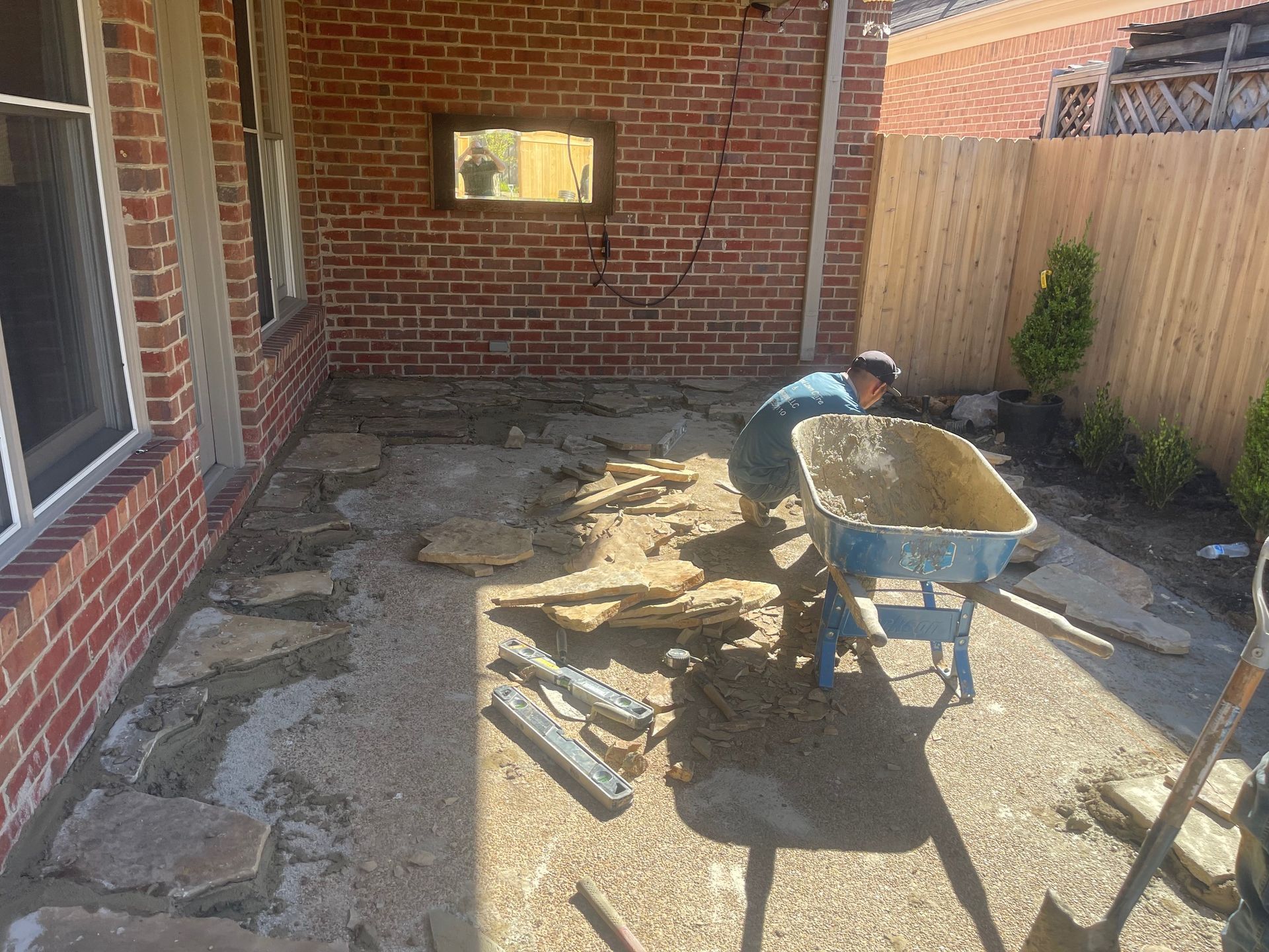 Person laying stones on patio with wheelbarrow; brick wall, wood fence, and sunlight present.