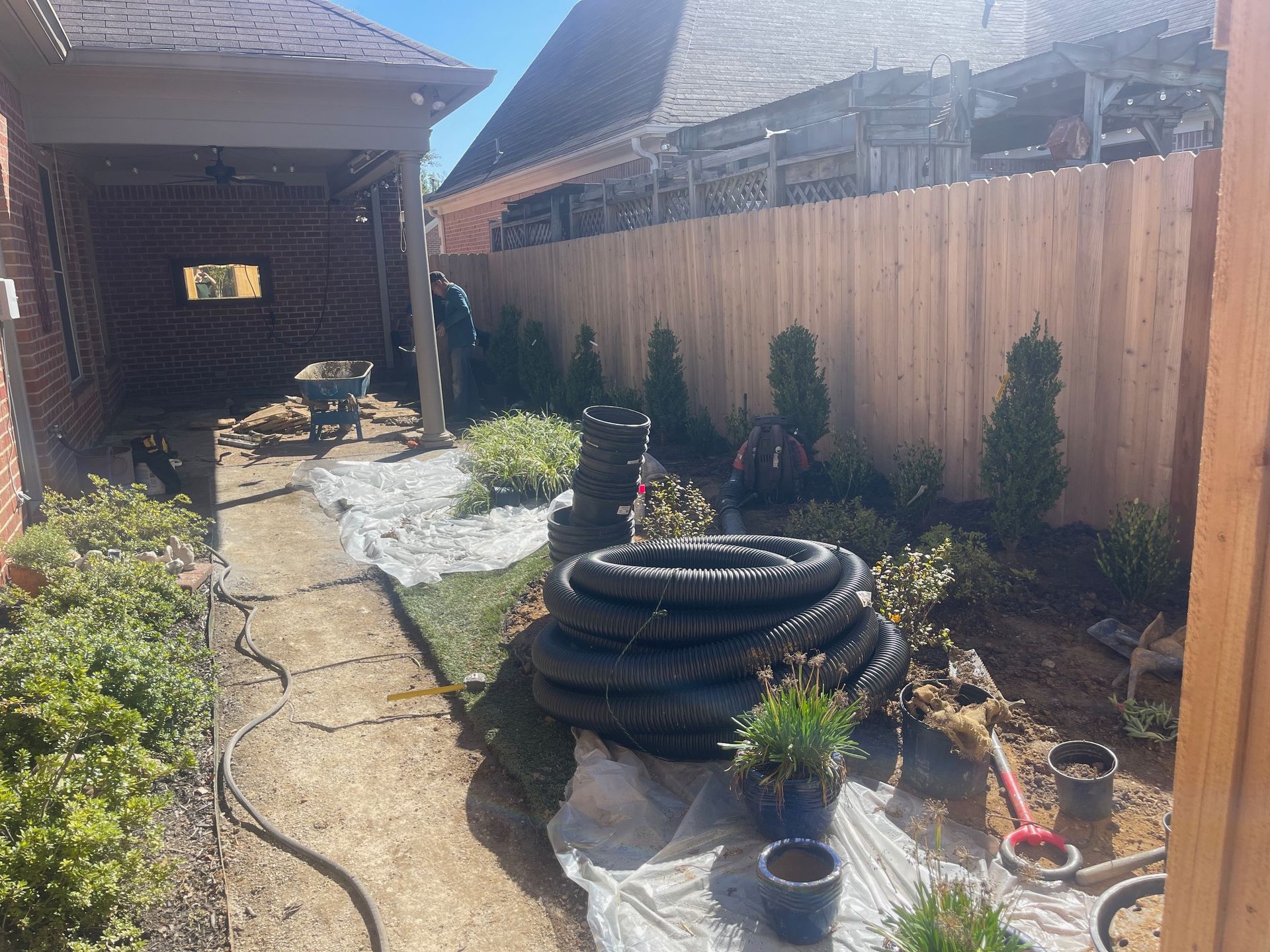 Landscaping work in progress: backyard with fence, plants, rolled tubes, and a brick structure.
