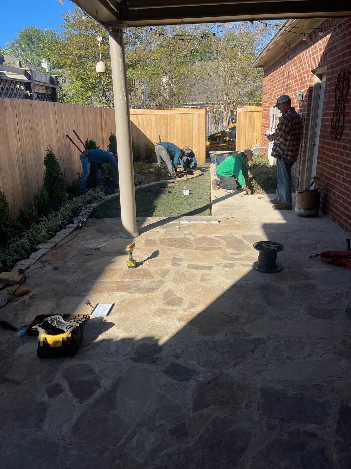 Workers installing landscaping near a patio and wooden fence on a sunny day.