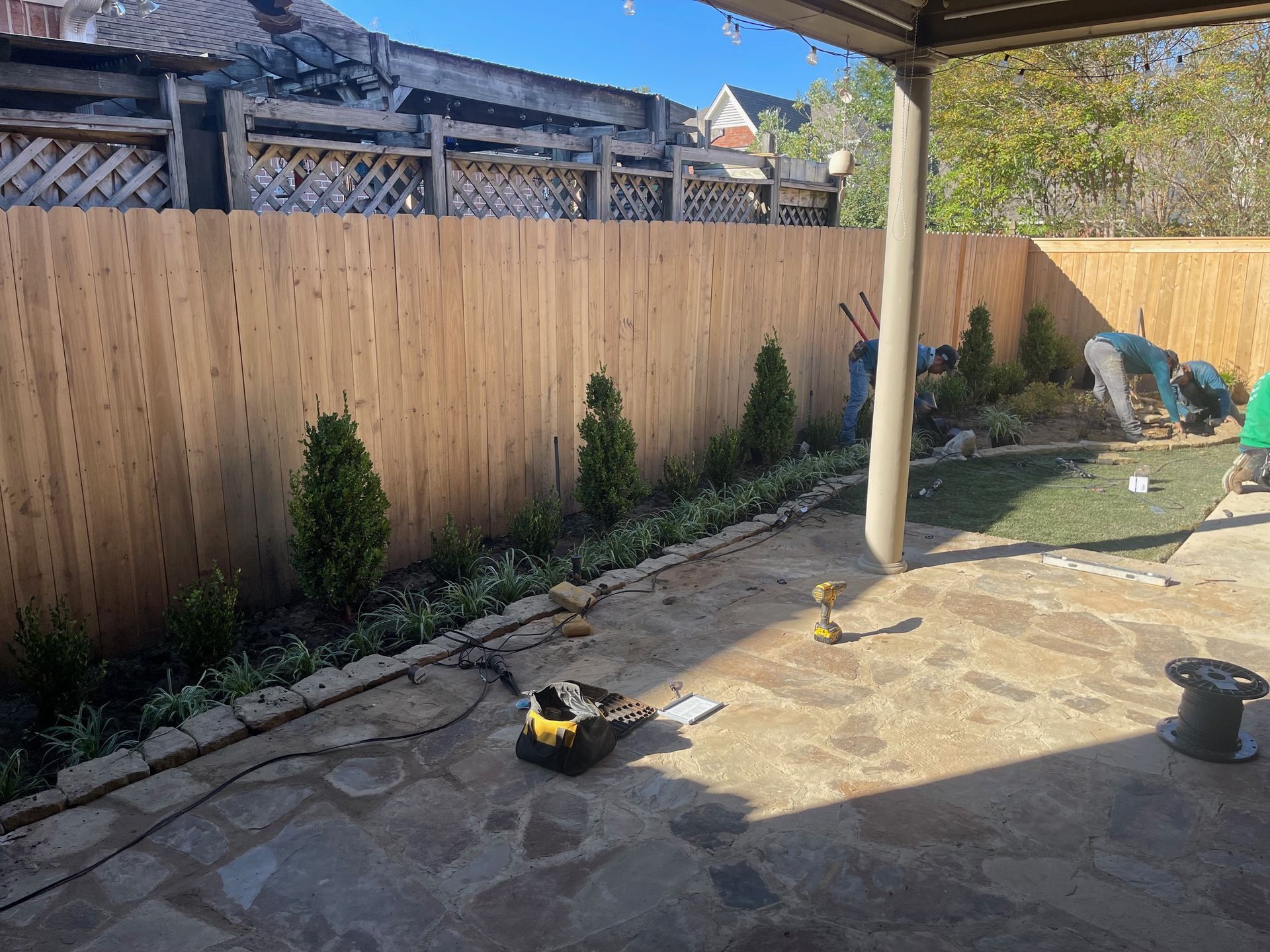 Backyard landscaping with new fence, stone patio, small trees, and workers.