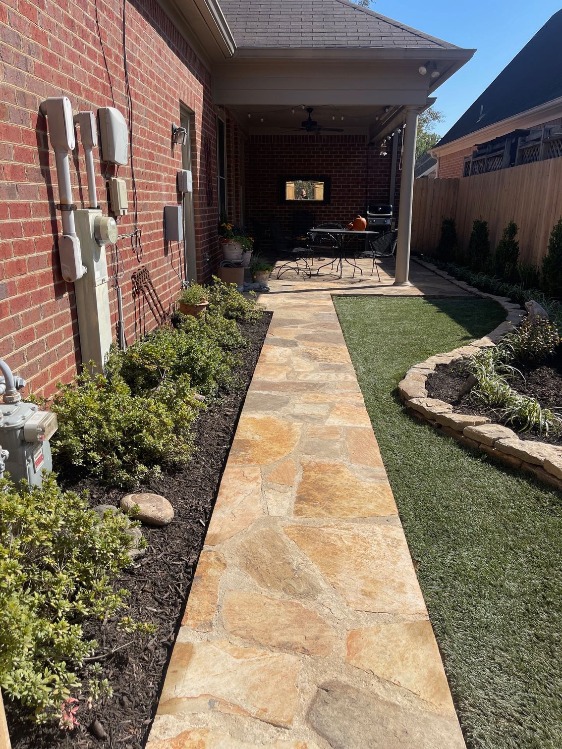 Stone path leads to a covered patio with greenery and lawn on either side; brick wall on left.