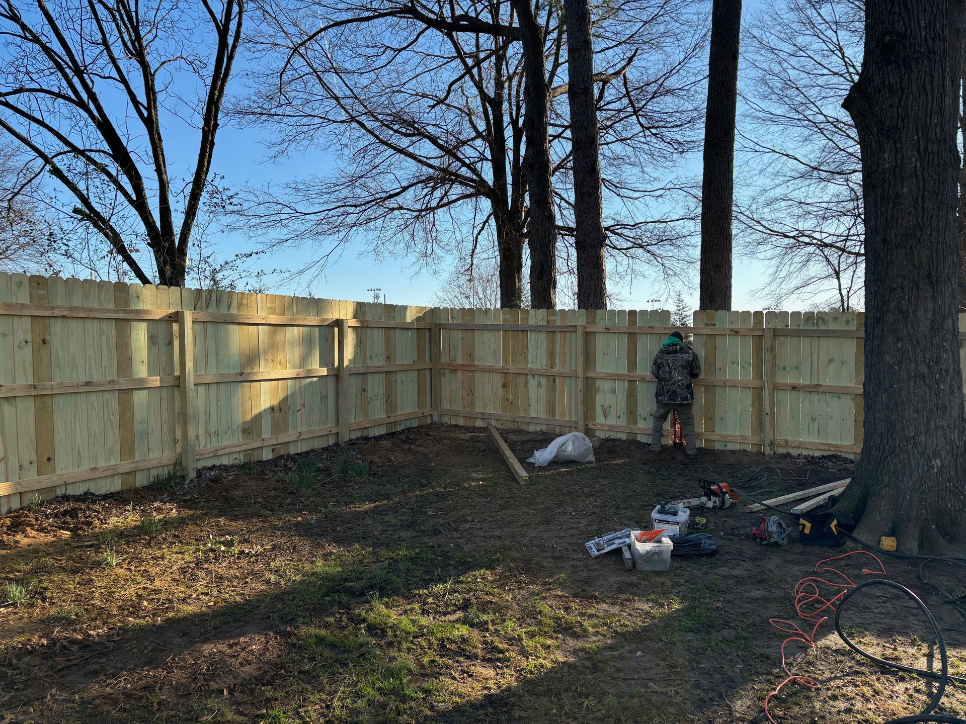 New wooden fence partially built in a backyard with a person working, surrounded by trees.