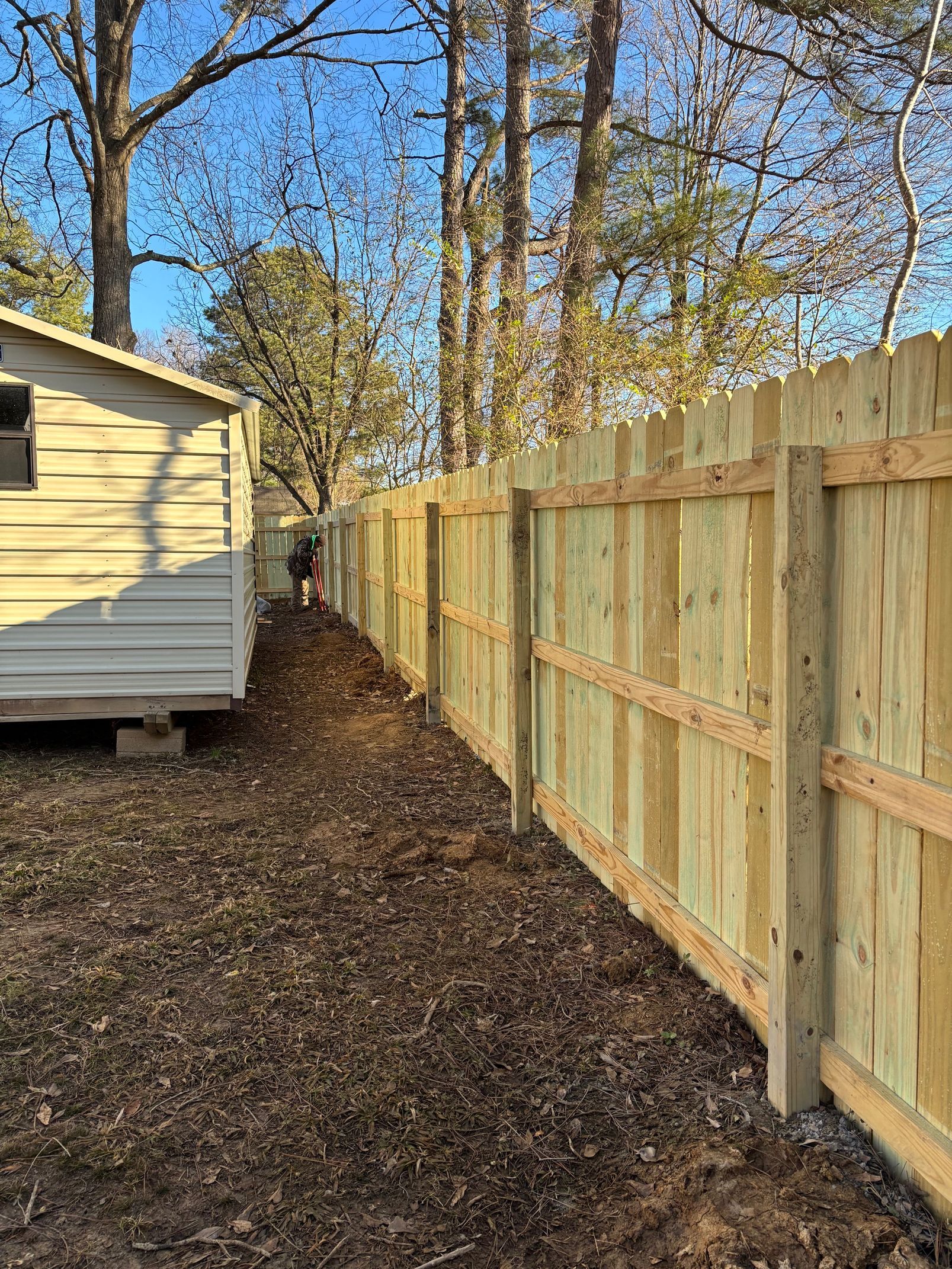 A wooden fence alongside a tan shed in a yard with bare soil and a few trees.