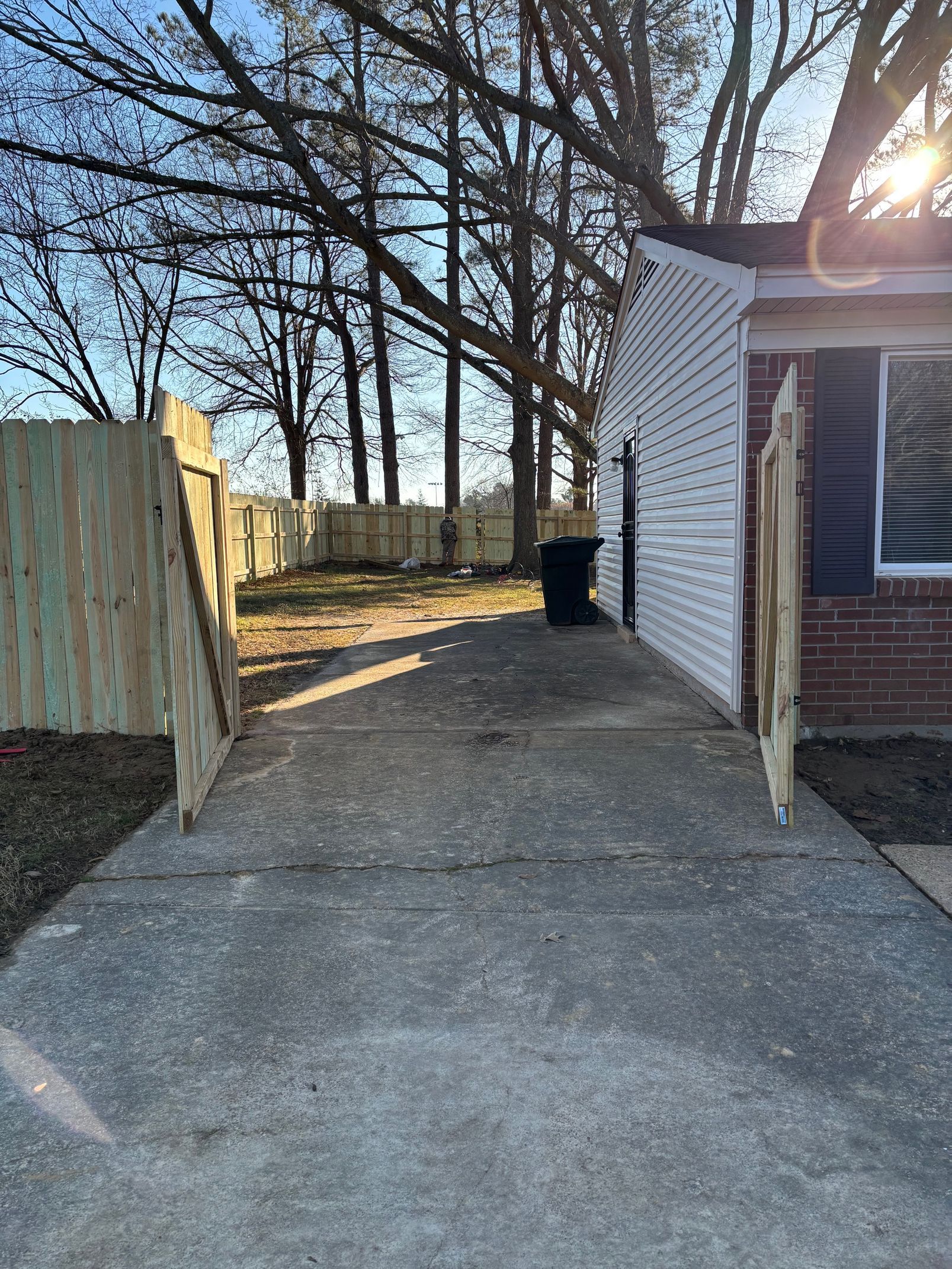 Concrete path leads to backyard between a white-sided building and wooden fence, sunny day.