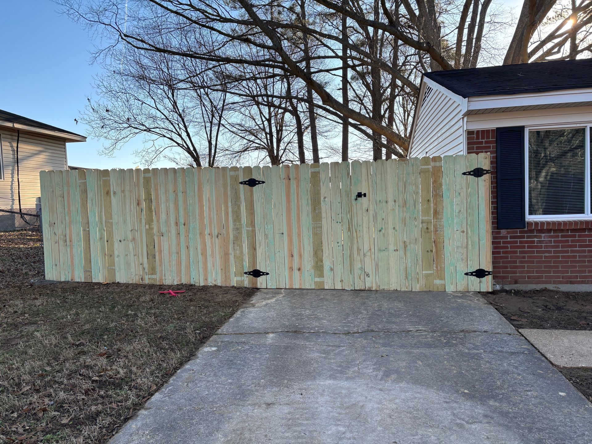 Wooden fence with black hinges in front of a brick house and driveway.