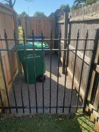 Green trash bin enclosed by fences and a black metal gate.