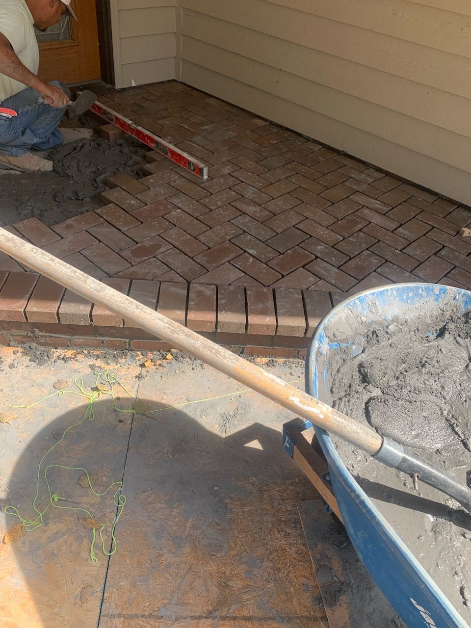 Person installing brick pavers in herringbone pattern on a porch. Mortar, wheelbarrow, and level visible.