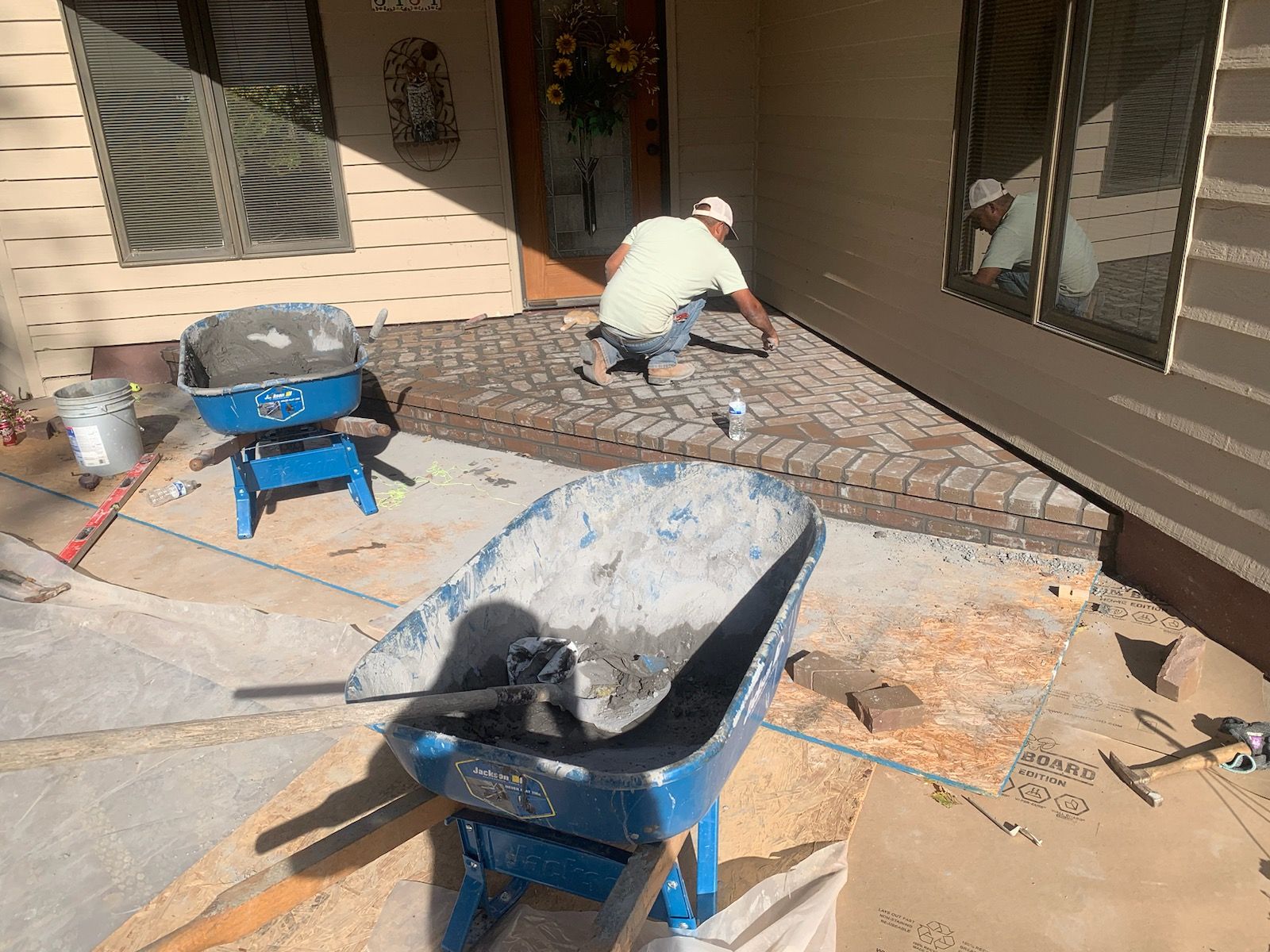 Man laying bricks on a porch. Blue wheelbarrows, tools, and brickwork are present. Sunlight illuminates the scene.