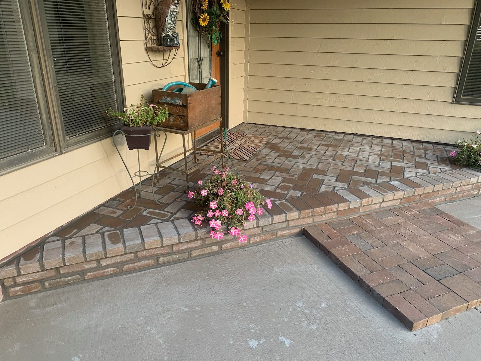 Brick patio with pink flowers in front of a house with a tan exterior.