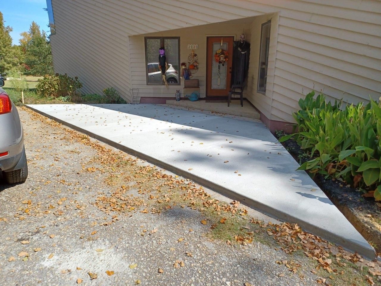 Concrete ramp leading to a house entrance, next to gravel driveway and greenery.