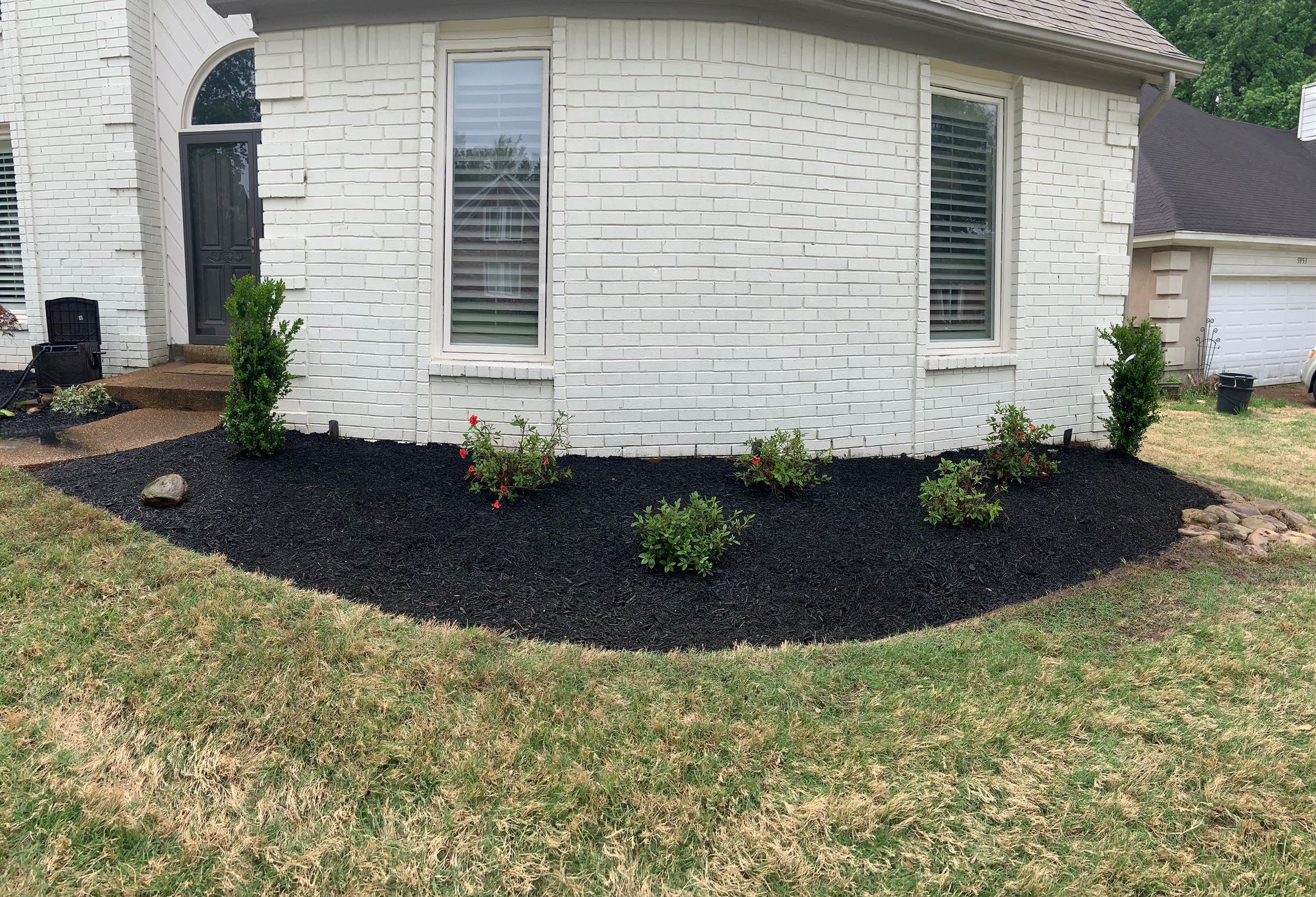 Black mulch bed with green bushes and small trees in front of a white brick house.