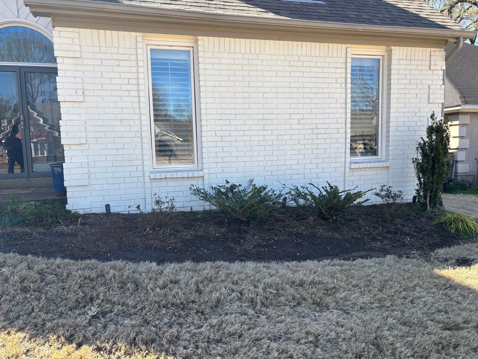 White brick house with two windows, brown mulch, and dry grass.