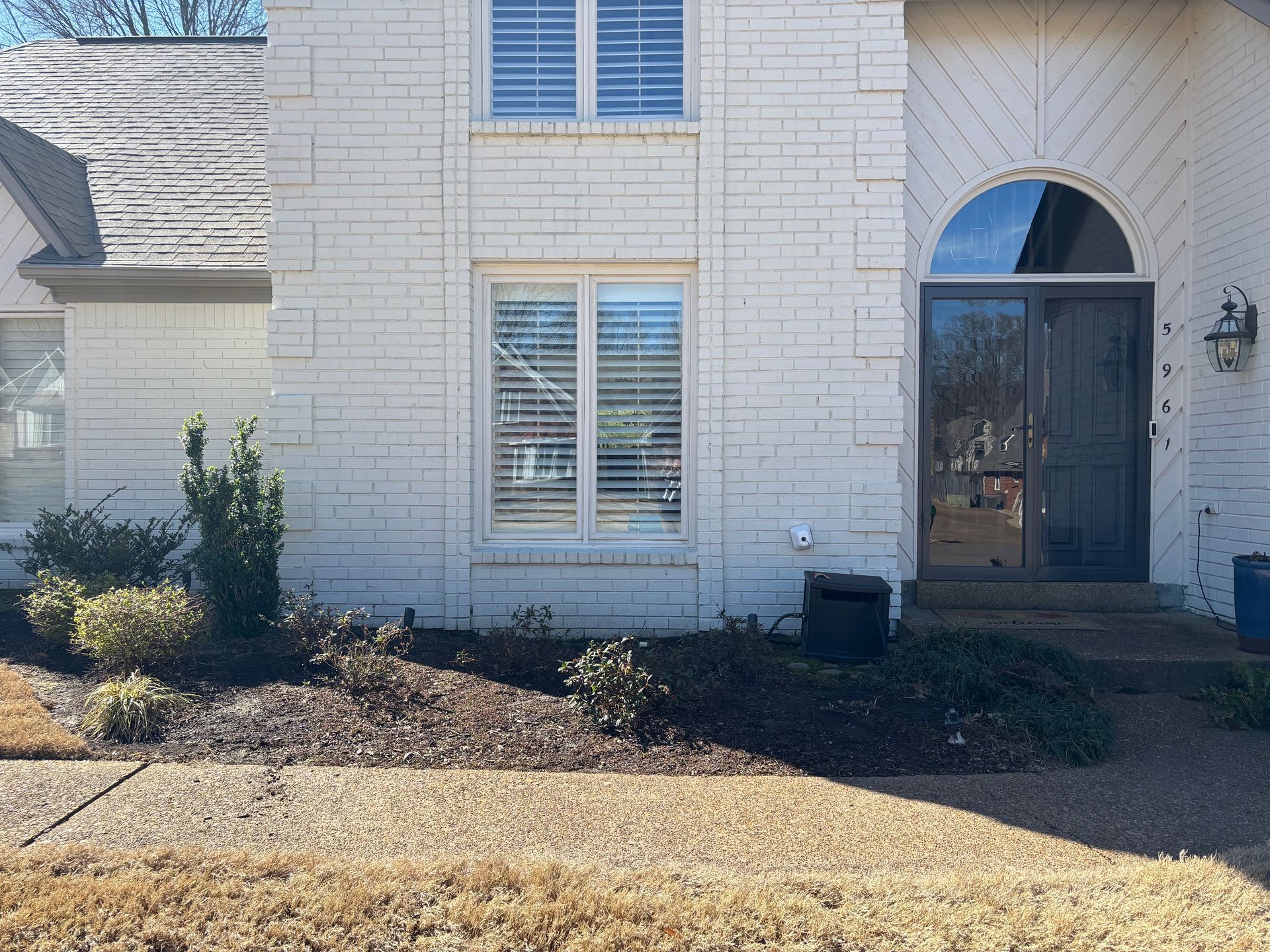 Off-white brick house with a small front garden. The front door is dark with a curved top and two windows with blinds.