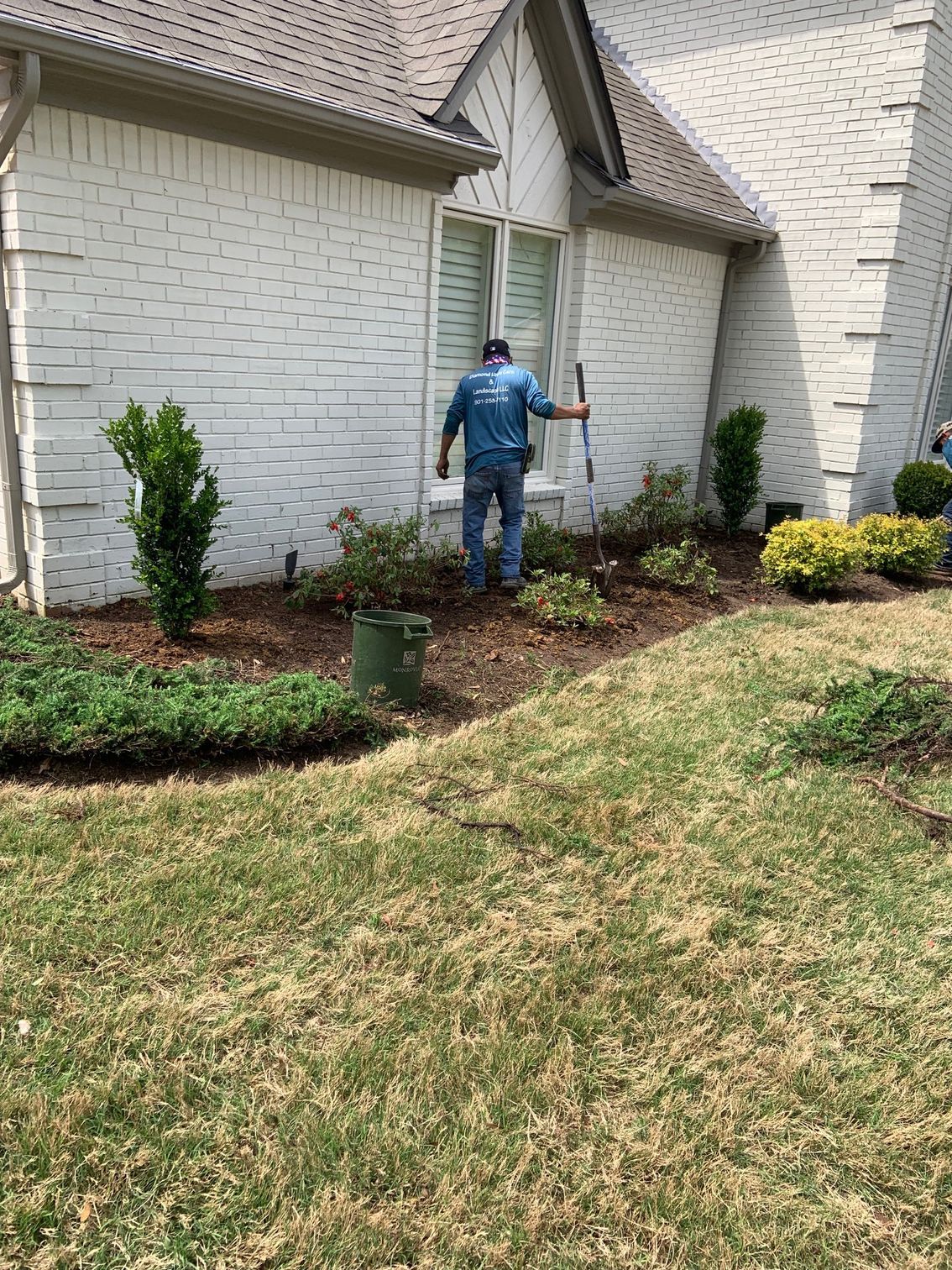 Person gardening in a flowerbed near a white brick house.