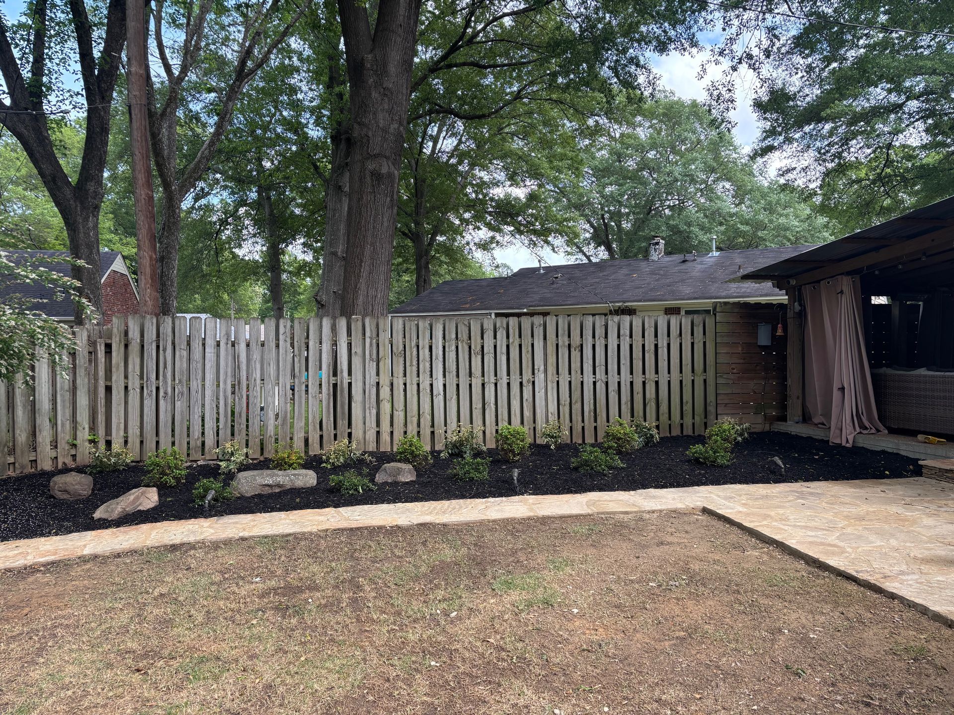 Backyard with a wooden fence, freshly mulched flowerbed, and trees.