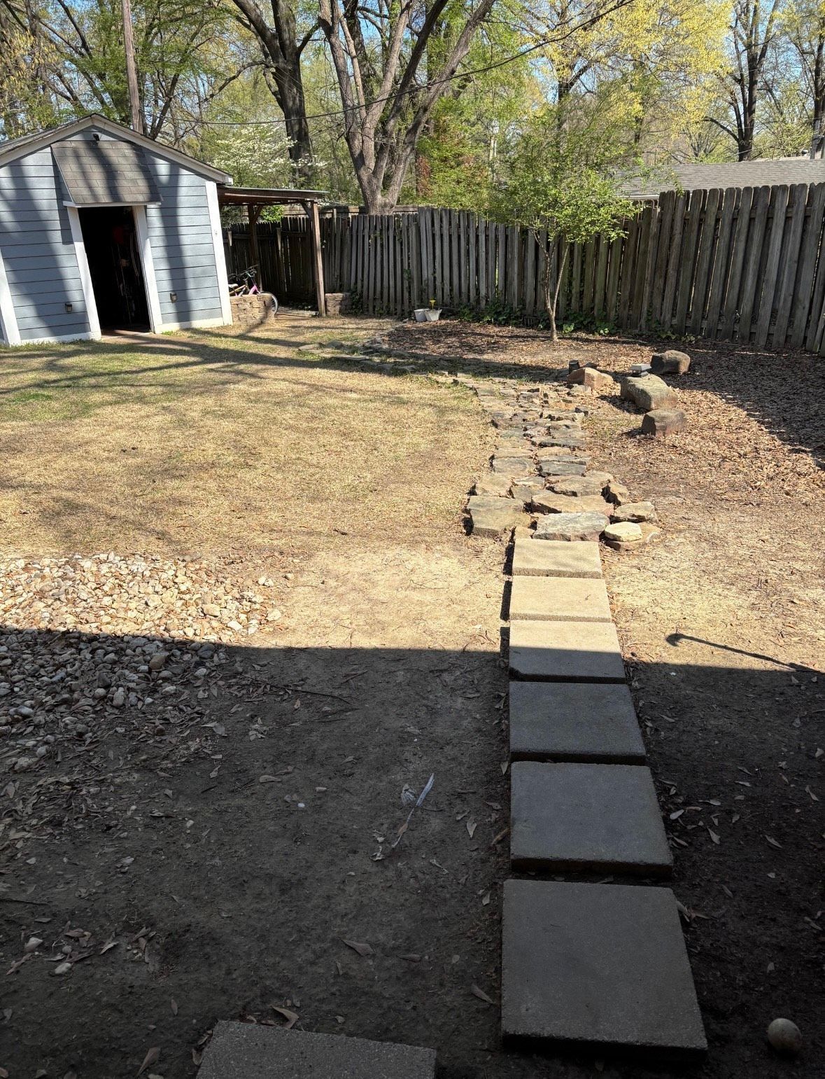 Backyard with stone path, shed, and wooden fence. Sunny day, brown grass, trees.